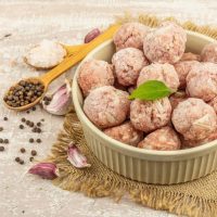 Frozen meatballs with spices, basil leaves, and garlic cloves in a ceramic bowl