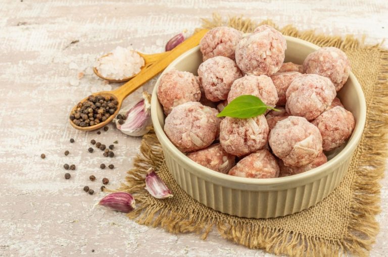Frozen meatballs with spices, basil leaves, and garlic cloves in a ceramic bowl