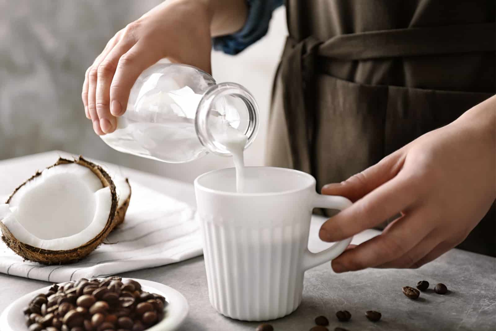 Woman pouring coconut milk into cup of coffee on table