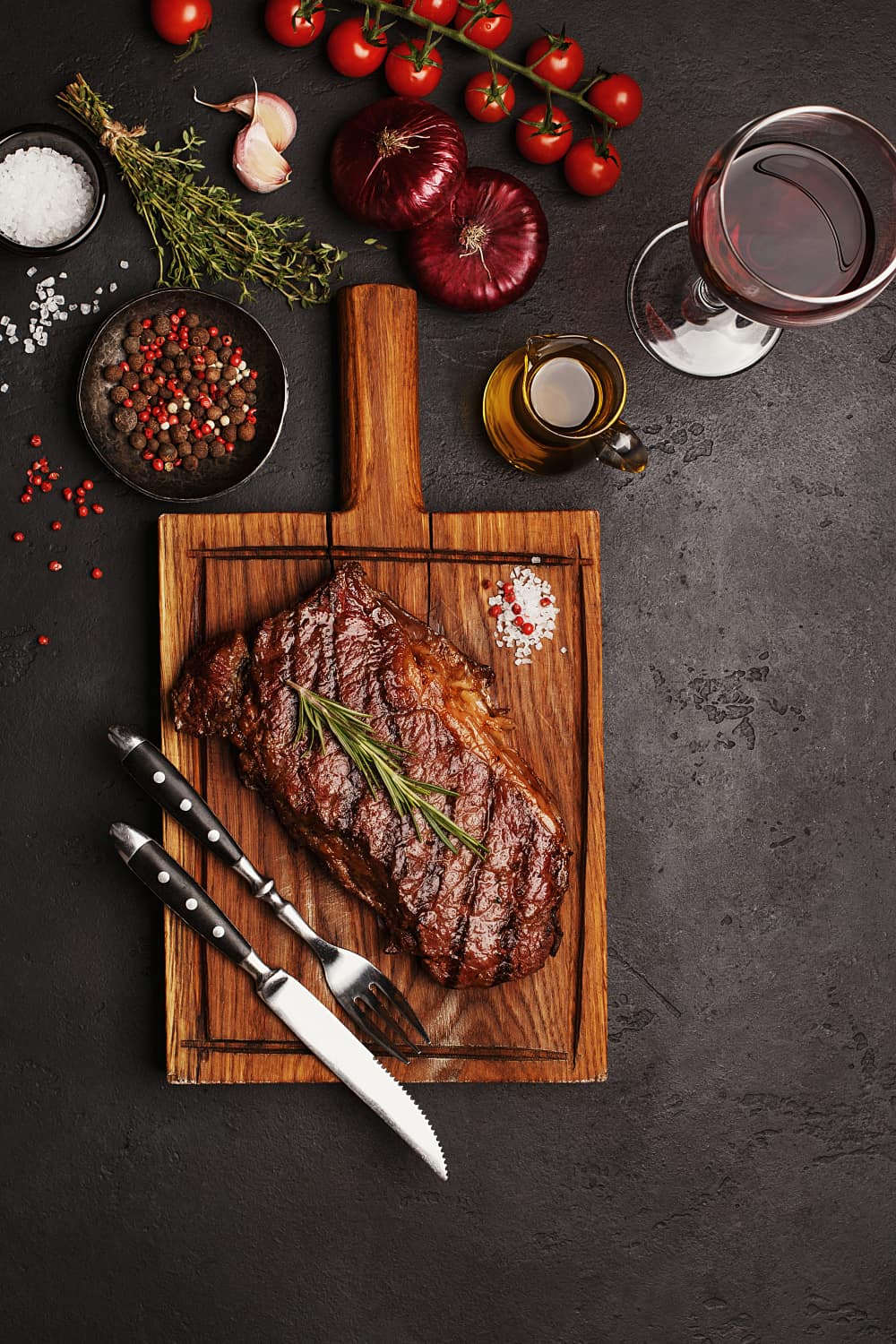 Striploin beef steak on wooden board with glass of wine, vegetables, herbs and spices on dark stone background