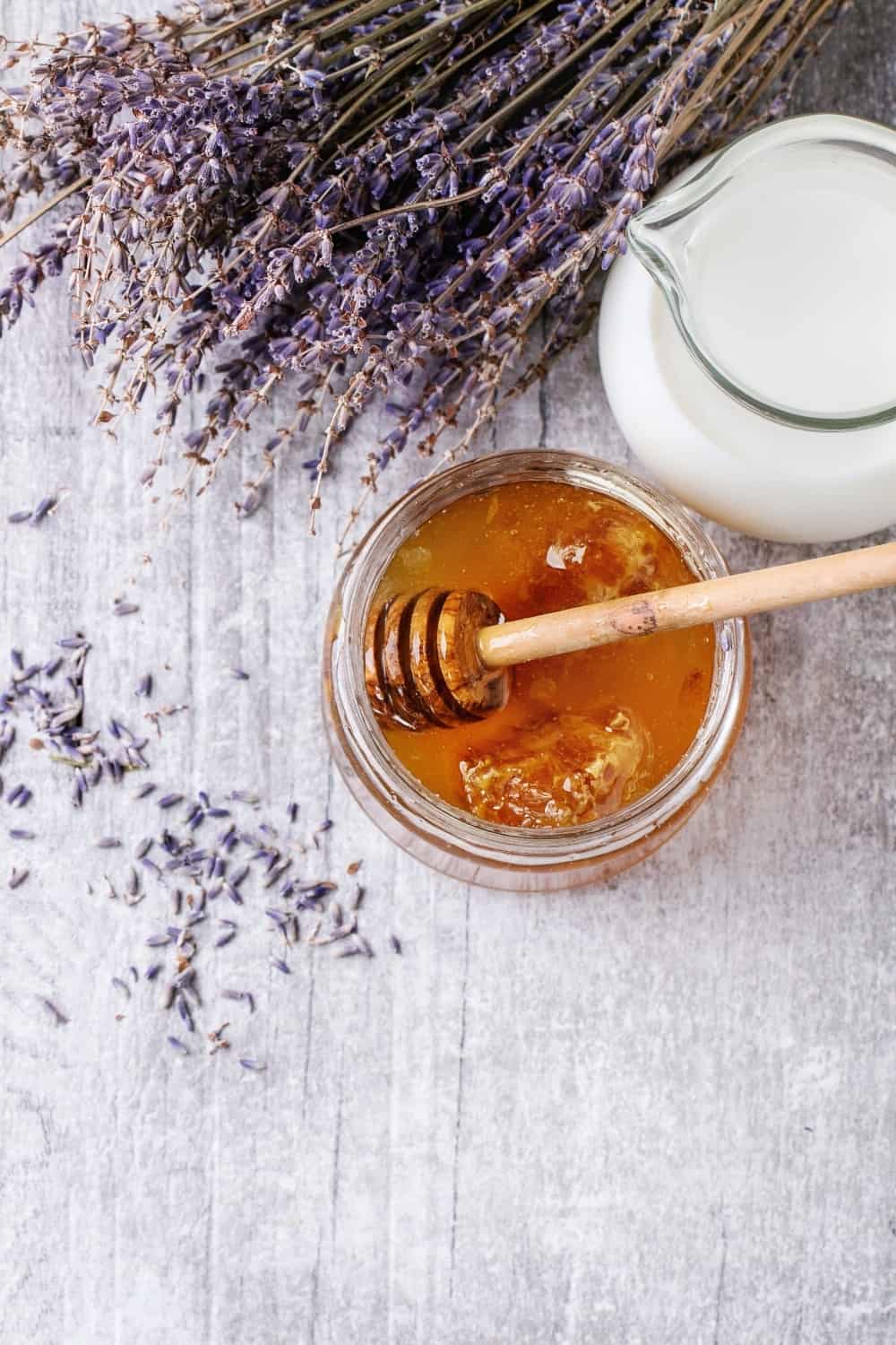 Tarro de cristal abierto de miel líquida con panal y cazo de miel dentro, jarra de cristal de leche y ramo de lavanda seca.