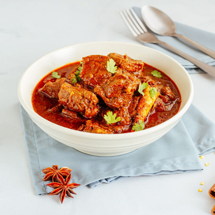 Caril de borrego Vindaloo de borrego numa tigela sobre fundo branco com colher e garfo.